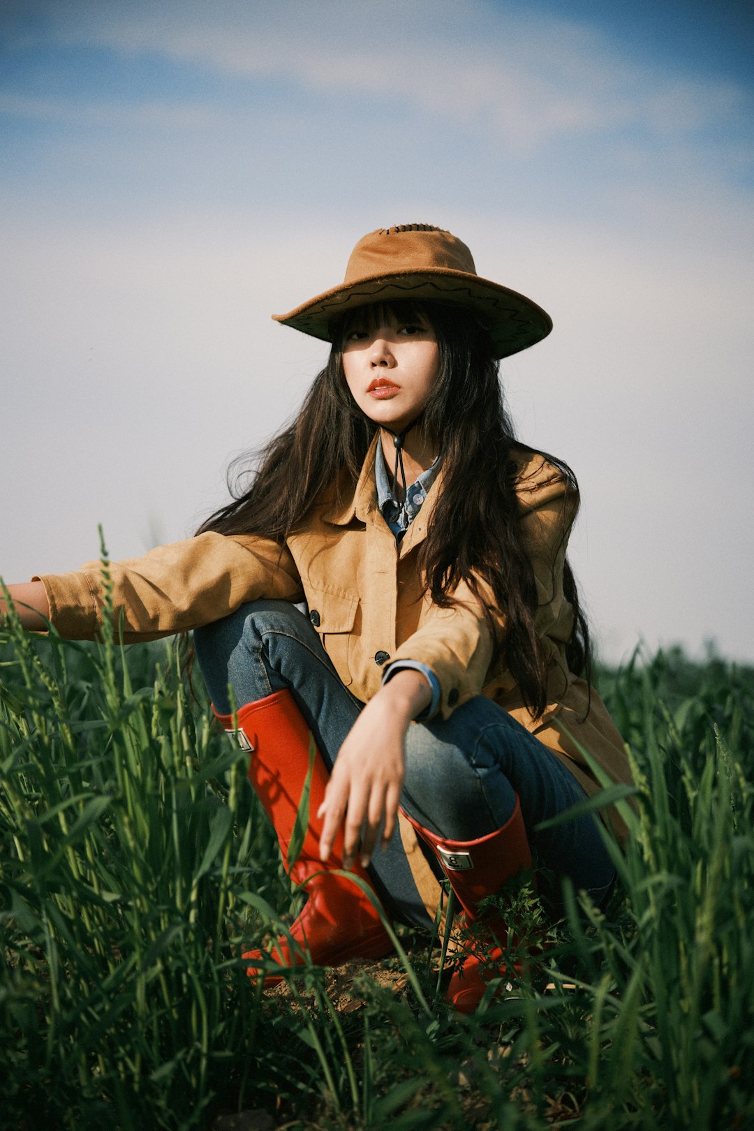 Woman in hat and boots squats in a green field.